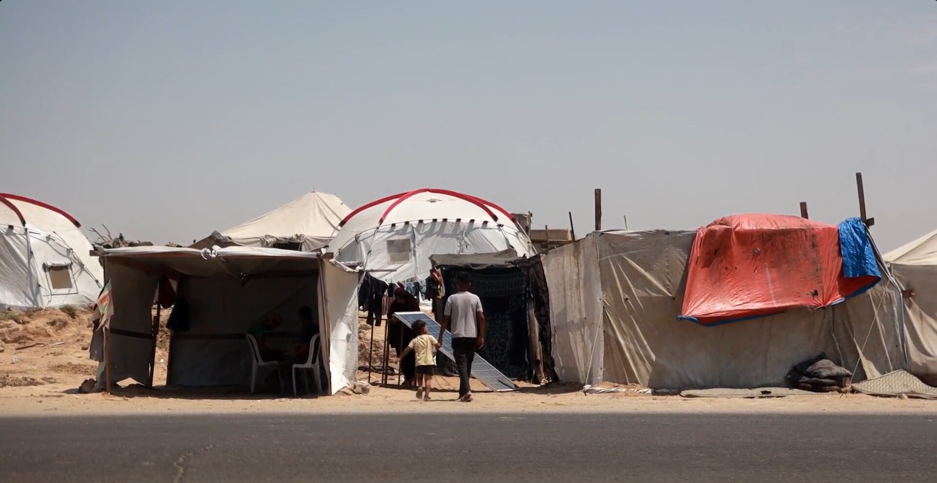 A boy walking hand in hand with a smaller boy near a sea of tents