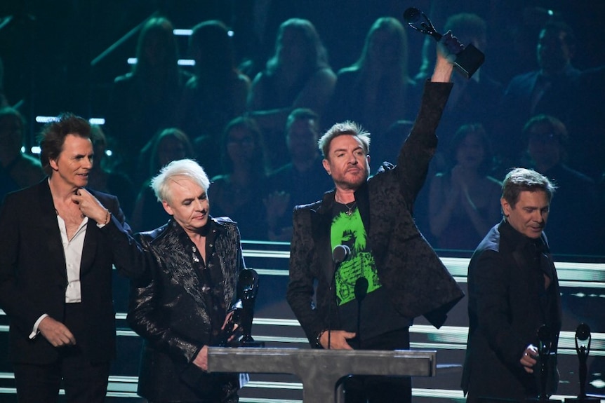 Four members of British band Duran Duran stand on stage. One holds award high above head. 
