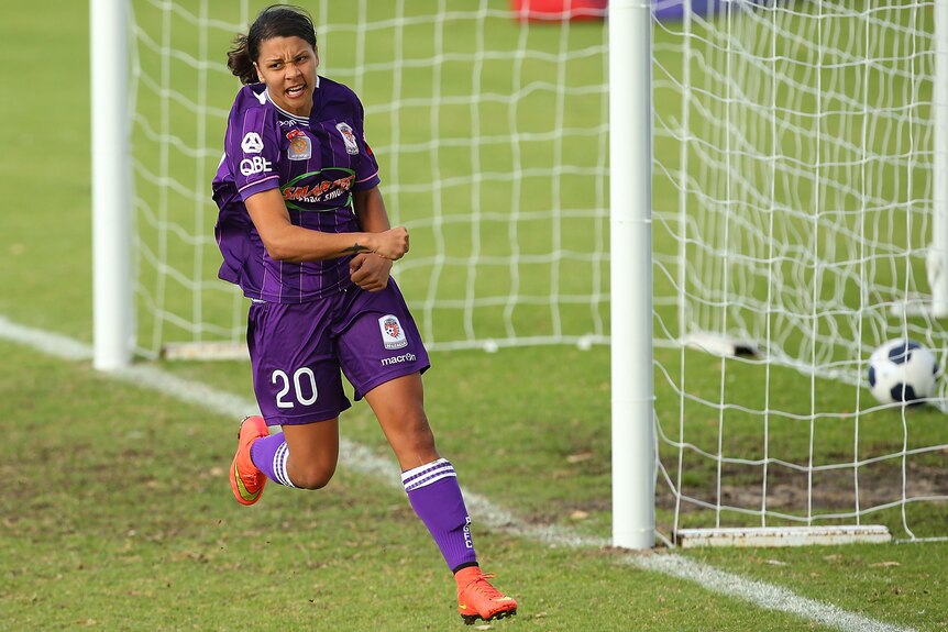 Sam Kerr runs past an empty goal net with a ball in the back with a grimace-smile on her face