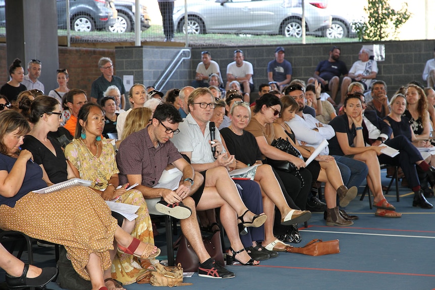 A man holds a microphone asking a question among a group of seated parents at a meeting