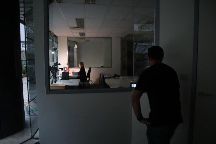 Woman sitting at desk with camera on tripod in front of her and cameraman in foreground watching through a window.