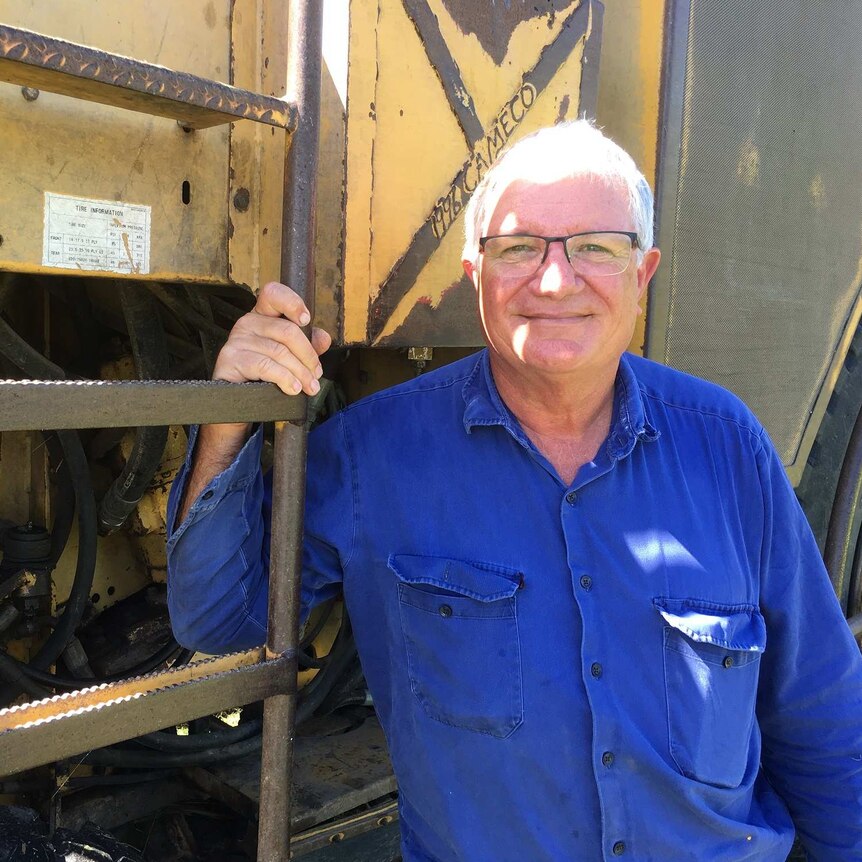 A man with white hair, wearing a blue shirt, standing in front of cane harvest machine, holding the attached steel ladder.