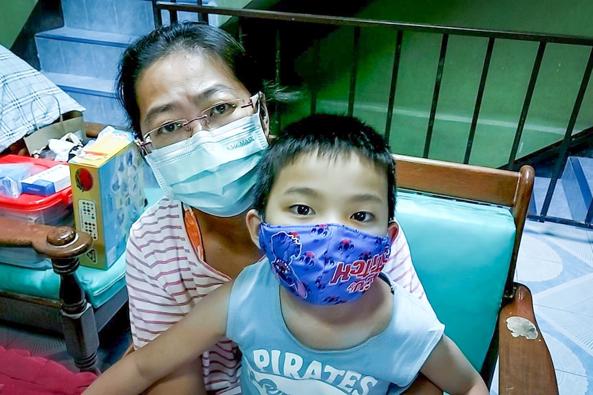 An older Thai woman in a face mask holds a small Thai boy, also in a mask, on her lap