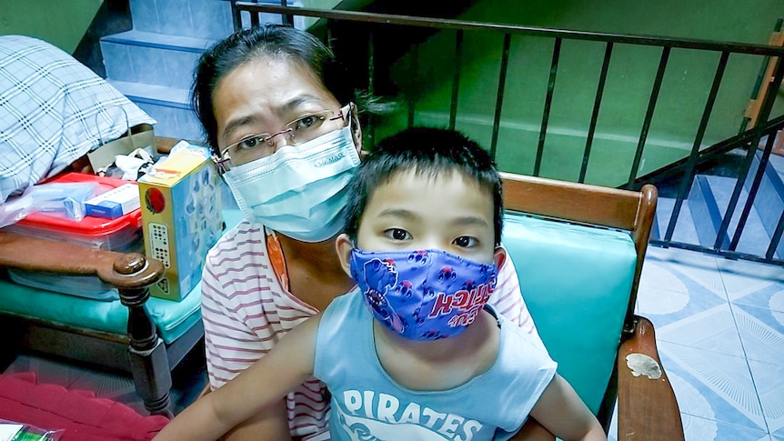 An older Thai woman in a face mask holds a small Thai boy, also in a mask, on her lap