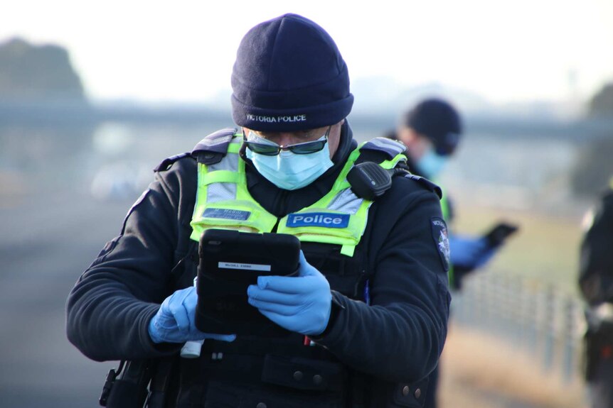 A police officer, wearing a mask and gloves, looks at a device in his hands on the side of the road.