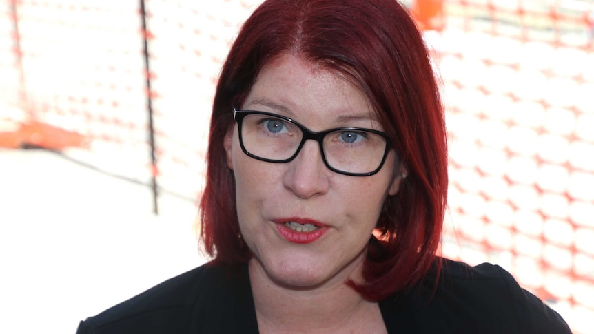 A close up of a red haired woman with glasses with sunshine and orange meshing in the background.