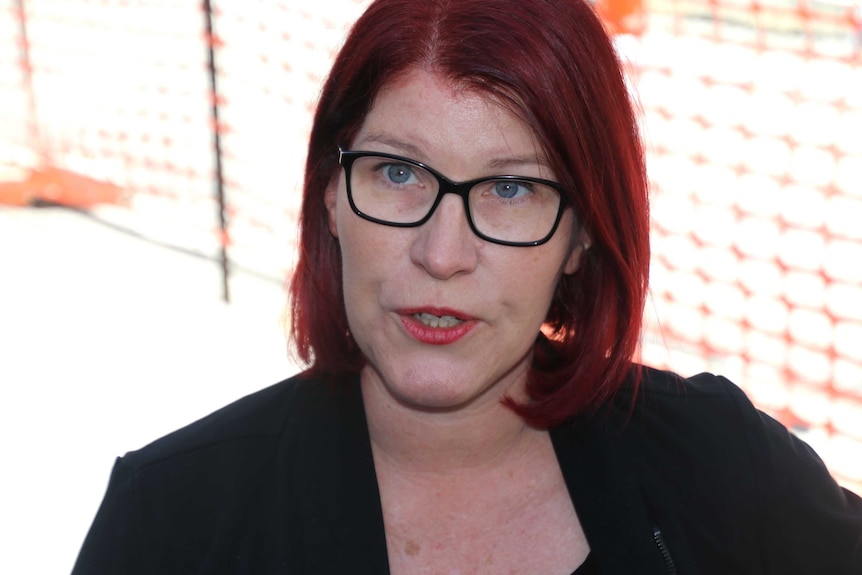 A close up of a red haired woman with glasses with sunshine and orange meshing in the background.