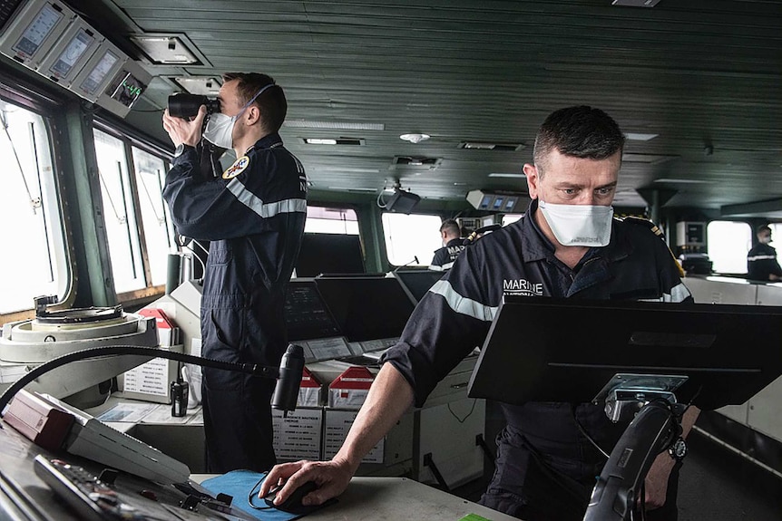 Two sailors wearing face masks, one looking through binoculars, work on a ship.