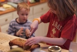 A girl cuts a slice of banana bread for her brother who is watching on