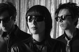 Black and white image of Yeah Yeah Yeahs wearing sunglasses, standing in a line, staring past the camera