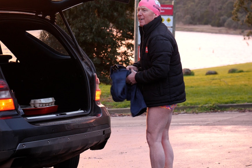 A man stands near an open car boot in front of a lake wearing a down jacket, pink bathers and swimming cap