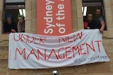 Students hang a white fabric sign with red writing on a building that reads "under new management".
