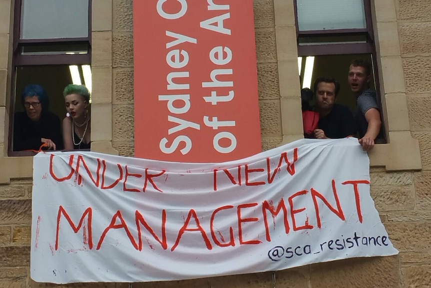 Students hang a white fabric sign with red writing on a building that reads "under new management".