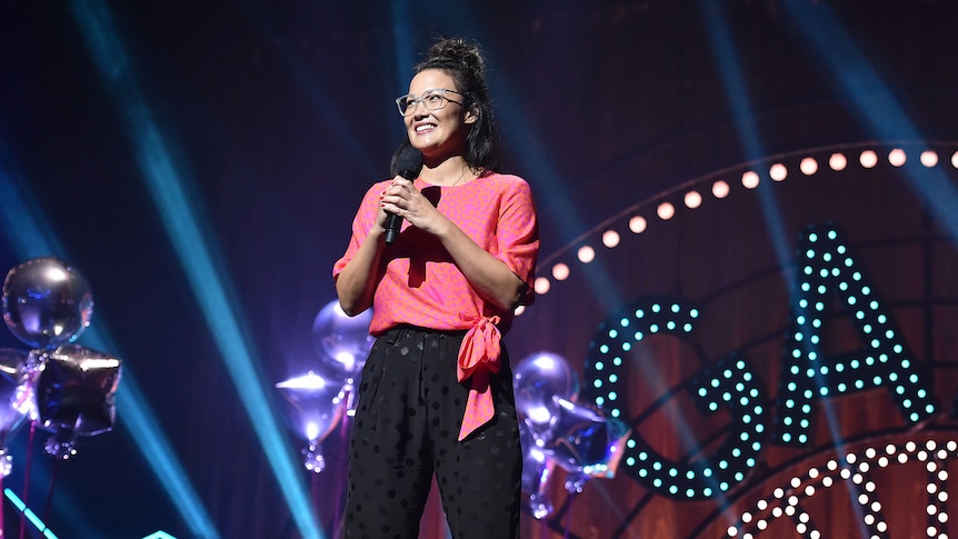 A TV still of Lizzy Hoo, a Chinese Australian woman in her late 30s, standing on stage holding a microphone.