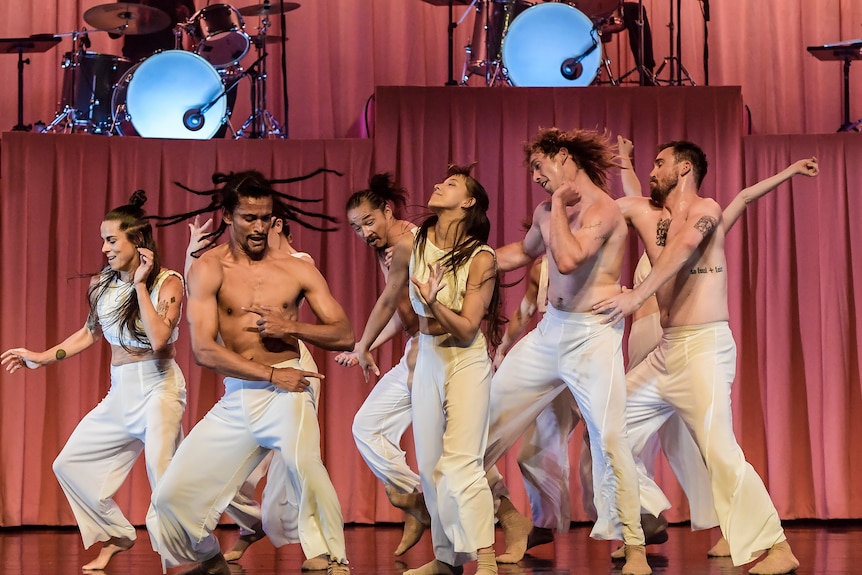 Nine dancers in white are mid-movement on a stage with a dusty pink curtain and drummers in the background.