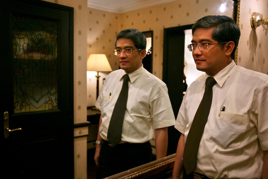 A man in a tie and short-sleeved shirt stands next to a mirror so his image appears twice