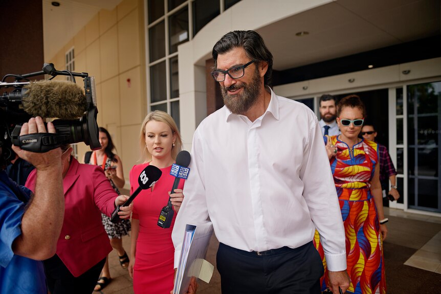 A white man with thick black facial hair exits court surrounded by TV reporters