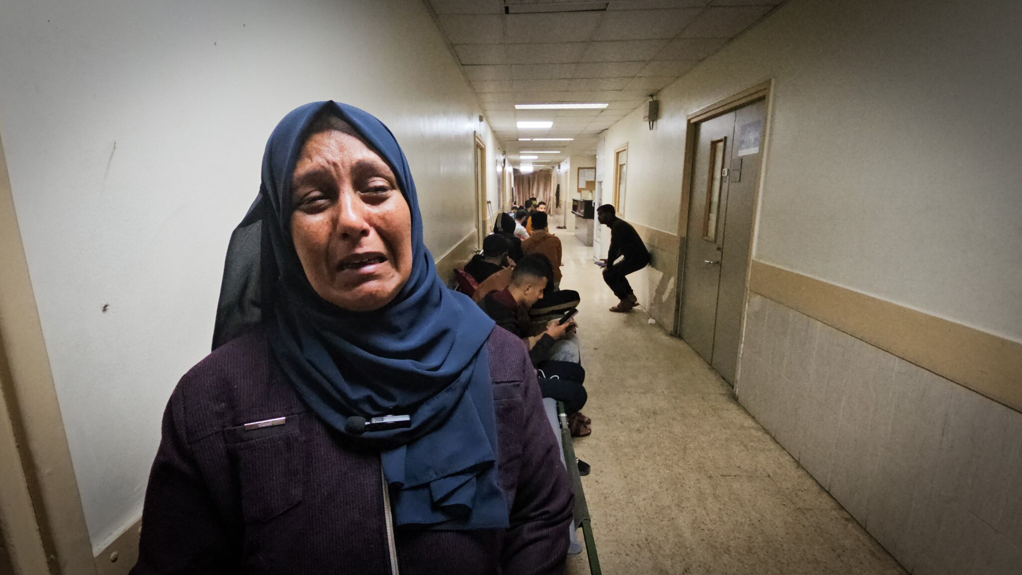 A woman wearing a dark blue hijab openly cries in a crowded hospital corridor.
