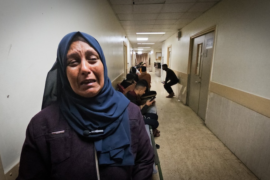 A woman wearing a dark blue hijab openly cries in a crowded hospital corridor.