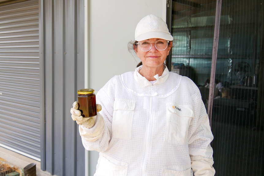 A woman wearing a white bee suit and cap holds a jar of honey.
