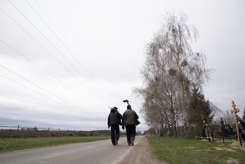 Two figures walk away from the camera down a country road with shovels resting on their shoulders