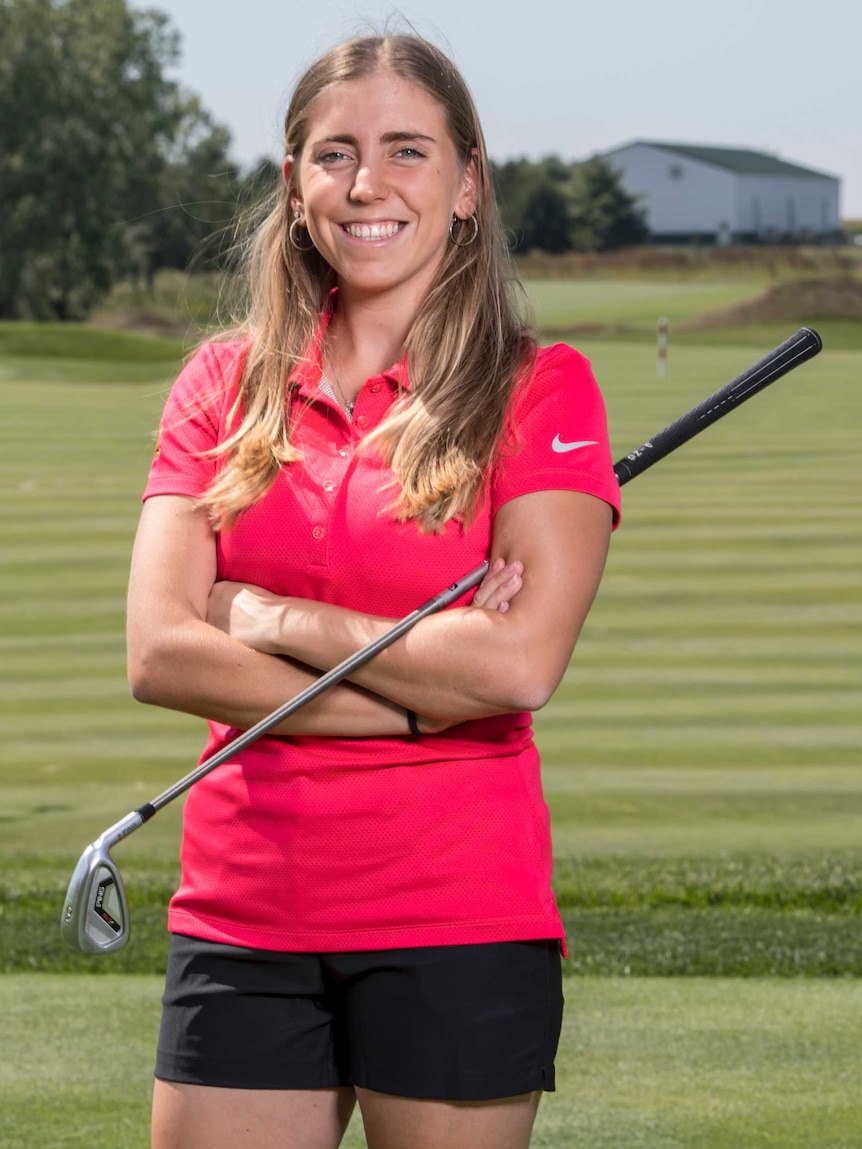 A female golfer smiles as she poses for a photo, club under her arm.