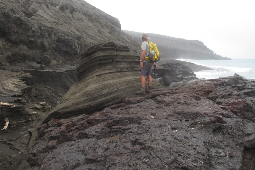 A man with a hat and backpack stands on volcanic rock and examines deposits left by a volcano near where he stands.