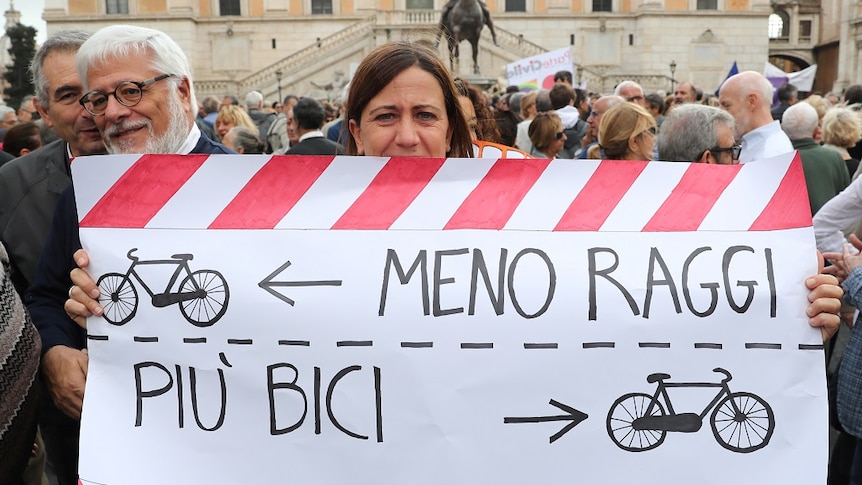 A woman holds a banner with text written in Italian, which translates to "Less Raggi, more bikes", and drawings of bikes.