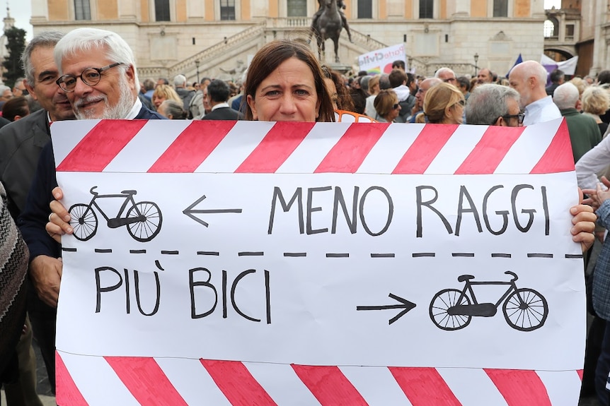 A woman holds a banner with text written in Italian, which translates to "Less Raggi, more bikes", and drawings of bikes.