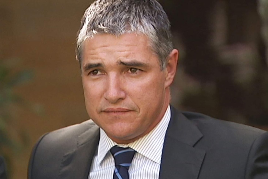 A man with short grey hair wearing a striped shirt, tie and dark suit.
