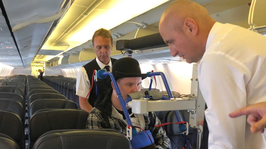 A man in a wheelchair wearing a bowler hat being pushed down the aisle of an aeroplane