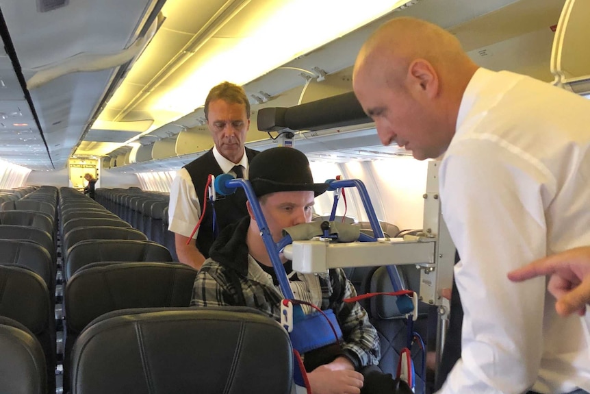 A man in a wheelchair wearing a bowler hat being pushed down the aisle of an aeroplane