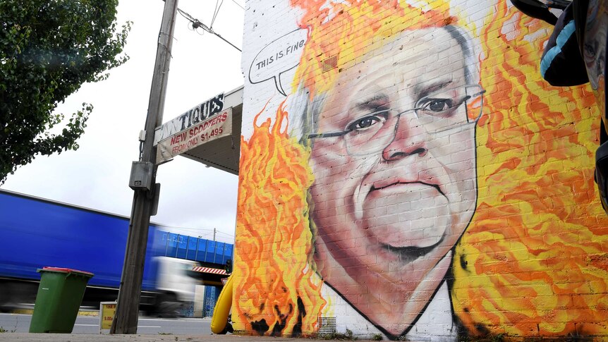 Scott Morrison is painted on a wall surrounded by flames, with a speech bubble that says 'this is fine'.