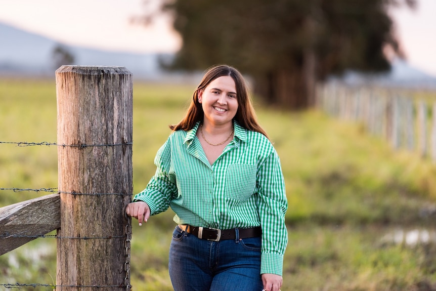 A woman in green checked shirt smiles as she stands next to a timber gatepost.