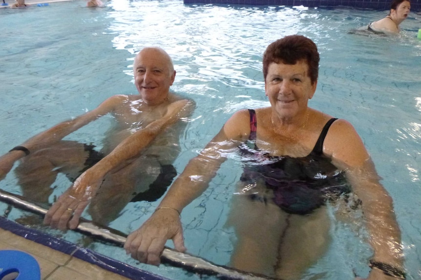 An older man and woman smile for a photo in an indoor pool, holding onto a side bar.