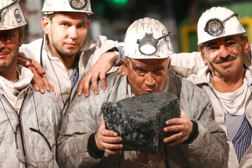 A miner kisses a piece of coal while his colleagues huddle around him.