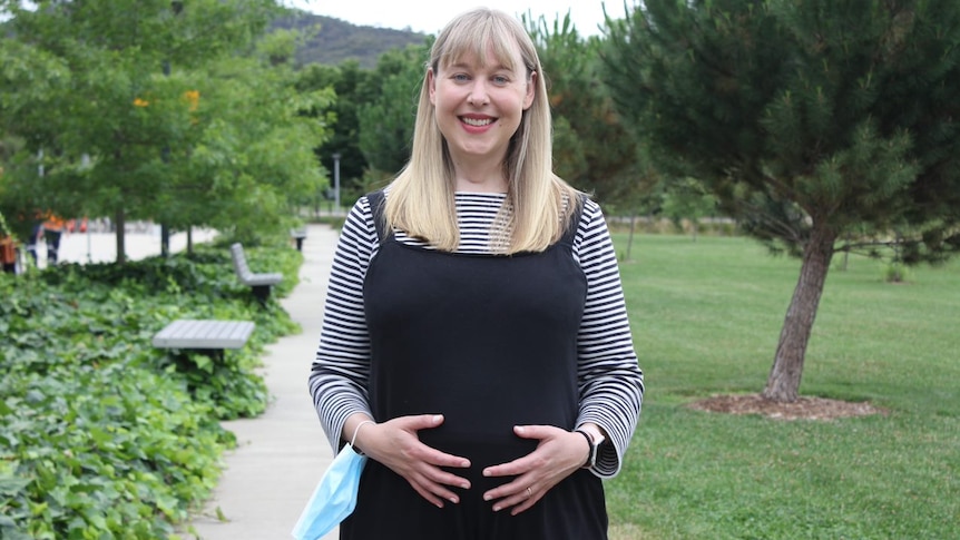 A pregnant woman in overalls holds her stomach.