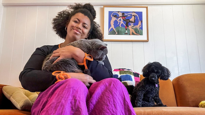 Yasmin smiles while holding Fig on a burnt orange couch with Molly on their right. A painting hangs behind them.