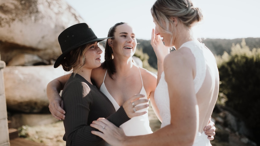 Two tearful brides embrace each other and a woman in a brown dress 