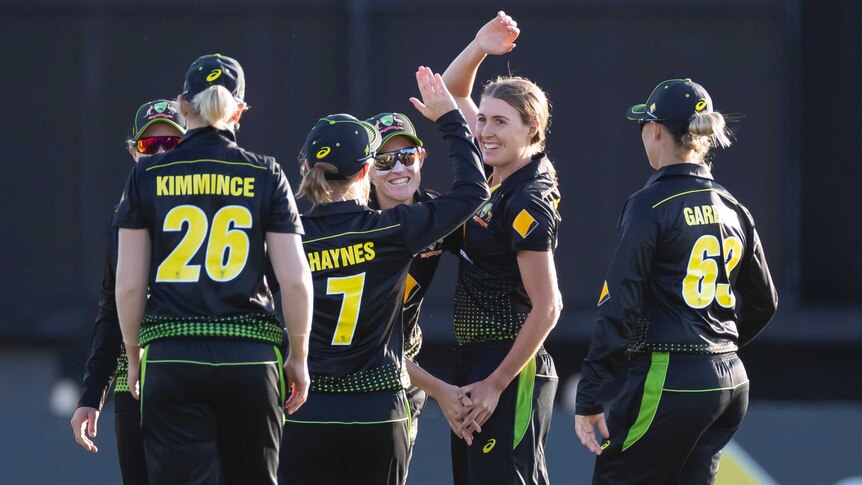A female cricketer raises her right arm as she celebrates with her teammates, who are surrounding her.