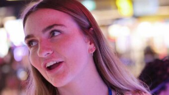 Keely Johnson looks to the left and smiles as she stands inside a bowling alley. She wears a tan jacket and leopard print top.