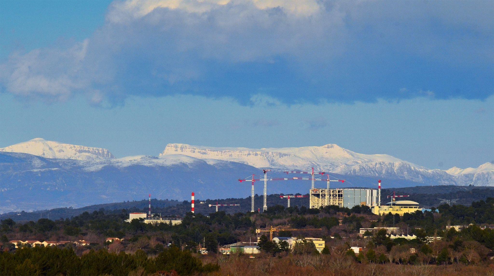 Tall cranes loom over large cement buildings, trees and forest surround the complex and snow-capped mountains loom behind it