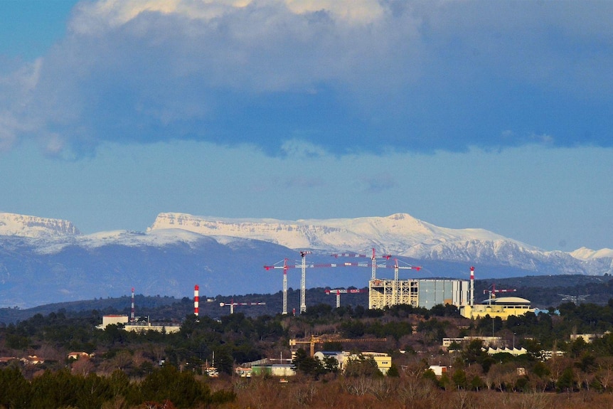 Tall cranes loom over large cement buildings, trees and forest surround the complex and snow-capped mountains loom behind it