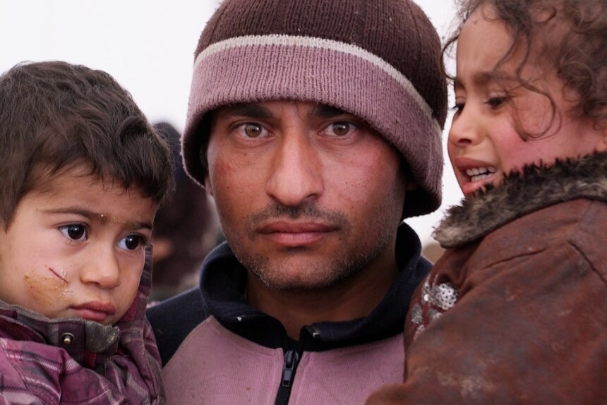 A man, staring at the camera, holds two children in a refugee camp