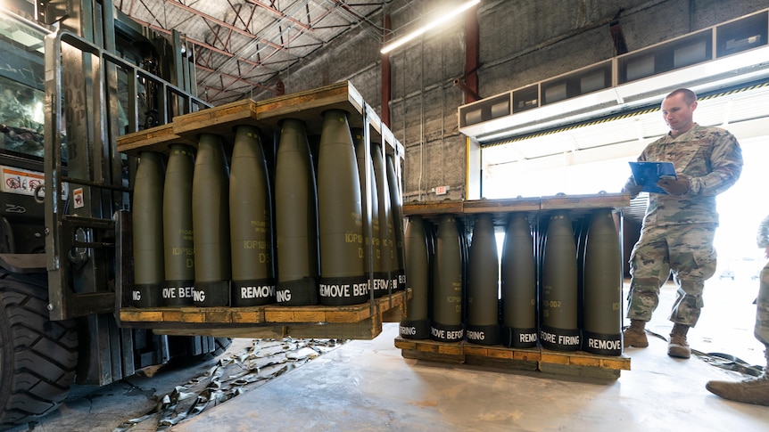 A US serviceman stands next to a pallet of artillery shells as another is moved by a forklift.