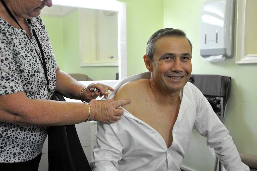 WA Health minister Roger Cook is given a needle by a nurse in a medical room.