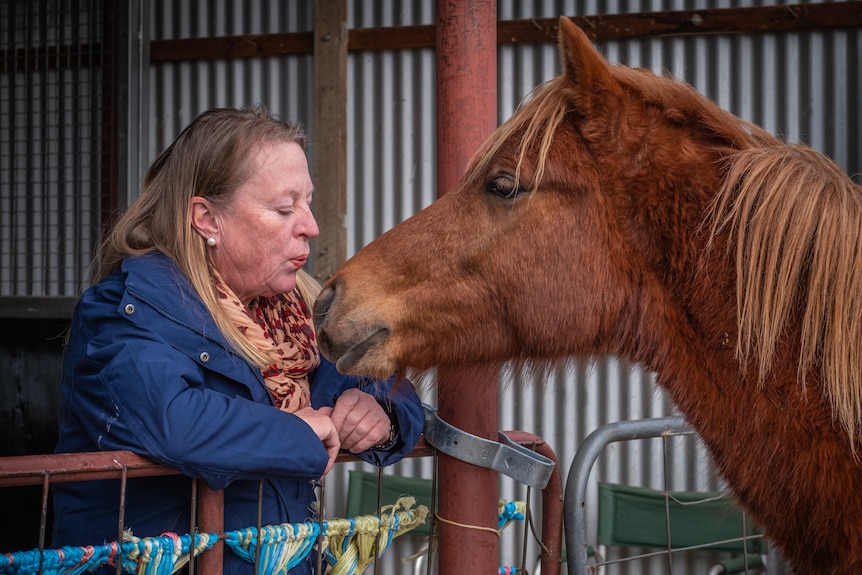 A woman with long blonde hair blows a kiss towards an orange-brown horse.