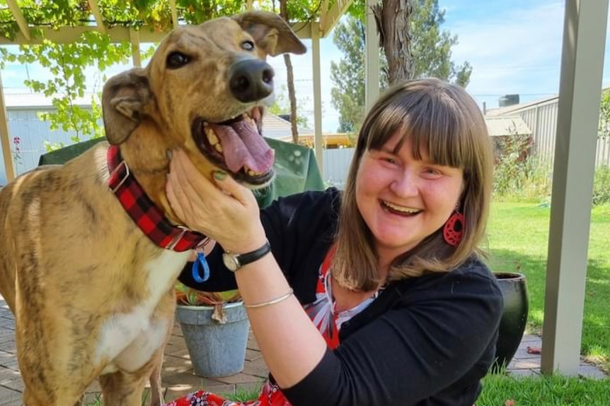 A woman with a huge grin on her face sits cross-legged on a lawn, playing with a happy dog.