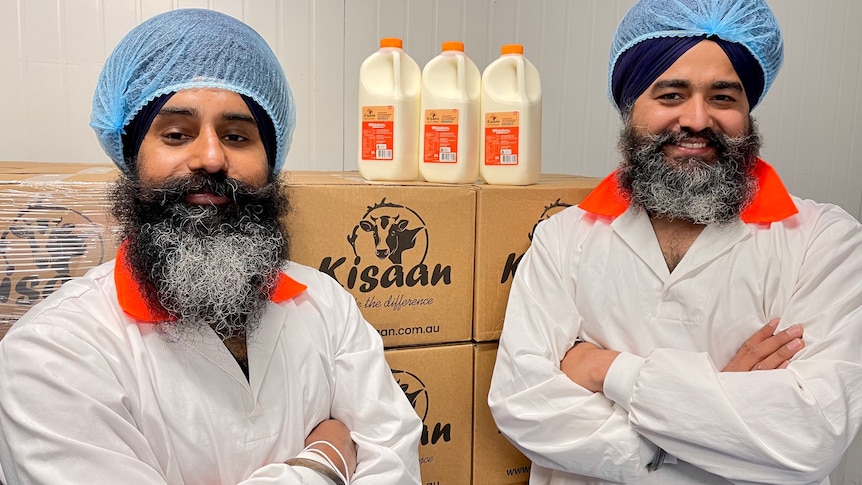 Two men stand in front of a stack of boxes, upon which three bottles of milk sit.