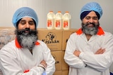 Two men stand in front of a stack of boxes, upon which three bottles of milk sit.